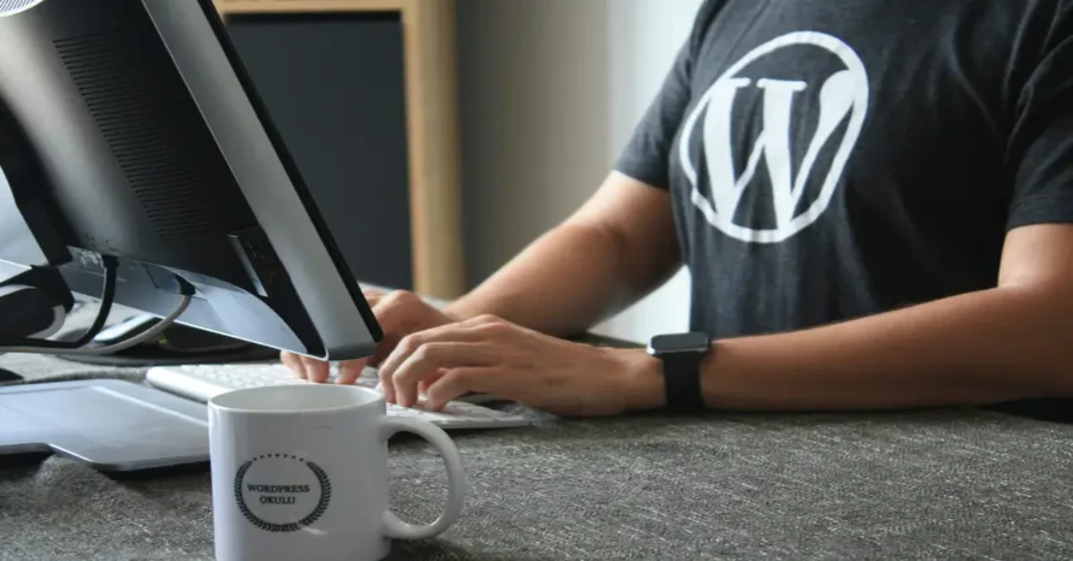 A person wearing a WordPress t-shirt is typing on a keyboard at a desk, likely fine-tuning the robots.txt directory. A WordPress-branded mug sits prominently in the foreground amid a sleek setup that includes both a computer monitor and a laptop.